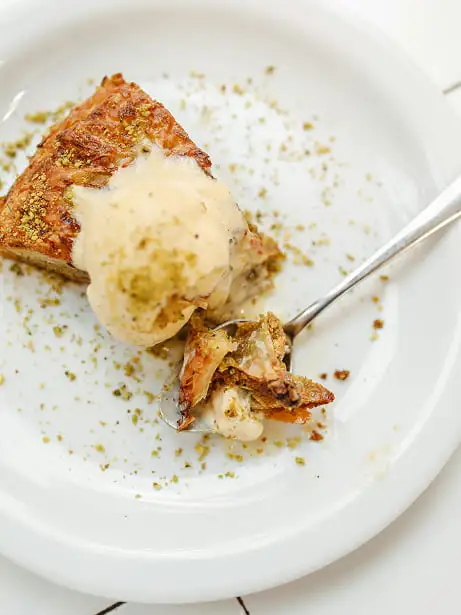 plato blanco con una cuchara plateada, dentro del plato hay una porción de bakclava de pistacho y una bola de helado de vainilla