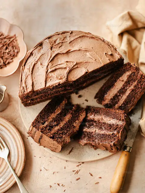 tarta de chocolate de tres pisos con crema de chocolate sobre una mesa que contiene un cuchillo, un trapo, un plato con cubierto y una pequeña jarrita