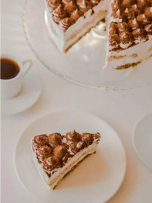 Peana arriba a la derecha de tartas con un tiramisu. A la izquierda una taza de café y en la parte inferior un plato con una ración de tiramisu